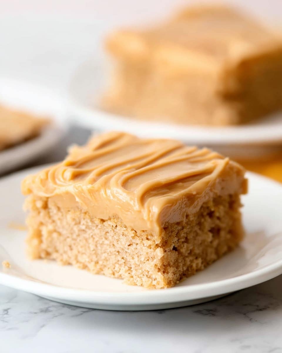 A close-up image of a single square piece of cake on a white plate, showing two layers: the bottom layer is a thick, light brown, soft and crumbly cake base, while the top layer is a smooth, creamy, slightly darker caramel-colored frosting with visible texture lines made by spreading. In the background, there is another similar cake piece on a white plate, slightly out of focus, all sitting on a white marbled surface. photo taken with an iphone --ar 4:5 --v 7