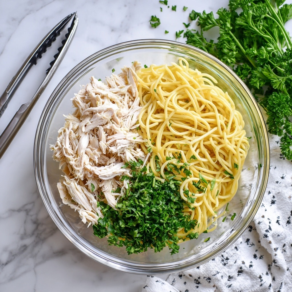 A clear glass bowl sits on a white marbled textured surface, filled with two main layers: shredded white chicken on the left side and cooked yellow spaghetti noodles on the right side. In the center, fresh bright green chopped parsley is spread as a garnish, adding a fresh pop of color on top of the chicken and noodles. To the left of the bowl, metal tongs rest on the surface. A bunch of curly parsley and a white cloth with small black patterns are placed in the top right corner. Photo taken with an iphone --ar 4:5 --v 7