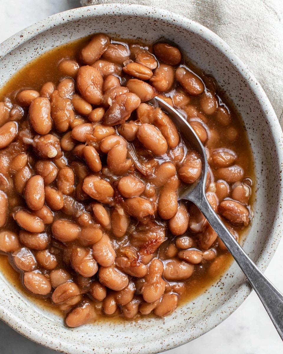 A close-up view of a bowl filled with cooked pinto beans in a light brown, slightly thick broth. The beans are soft and shiny, varying in warm brown shades with some lighter beans mixed in. A silver spoon is partially submerged in the bowl, scooping up a portion of the beans. The bowl itself is white with a speckled brown rim, sitting on a white marbled textured surface. The scene is brightly lit, emphasizing the rich, glossy texture of the beans and the broth. photo taken with an iphone --ar 4:5 --v 7