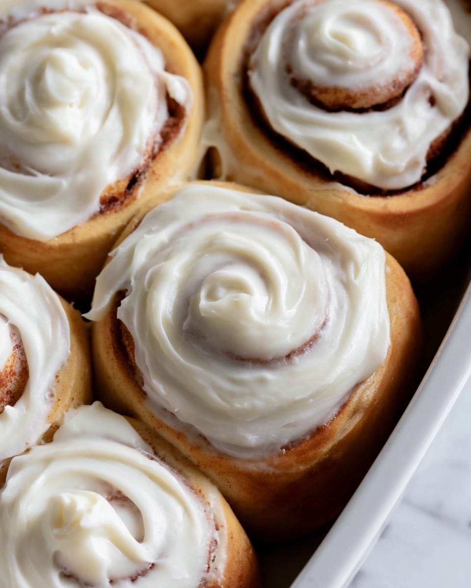 The image shows close-up of several cinnamon rolls inside a white baking dish. Each roll has soft brown dough with visible swirls of cinnamon filling inside. A thick layer of smooth white cream cheese frosting covers the top of each roll, with its creamy texture swirled gently to follow the shape of the curls. The background is a white marbled texture, which contrasts softly with the warm colors of the rolls. photo taken with an iphone --ar 4:5 --v 7