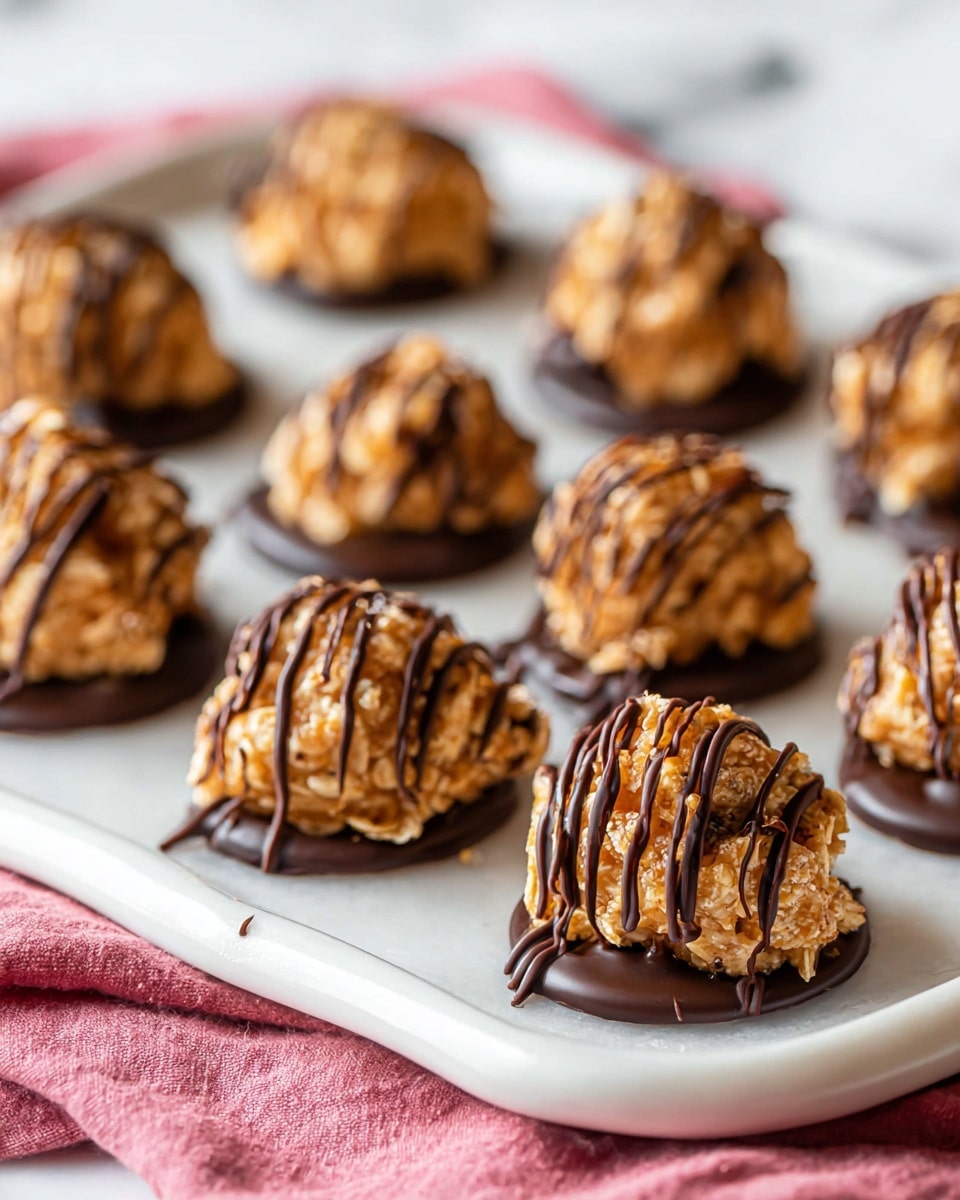 The image shows several small, round clusters of a light brown, textured mixture that looks like a no-bake cookie or candy, each with visible bits of nuts or oats. Each cluster sits on a thin, shiny dark chocolate base that spreads slightly around the bottom. Dark chocolate is drizzled on top of every cluster in fine lines creating a striped pattern. The clusters are arranged in rows on a large white tray with a smooth rim. The tray rests on a soft pink cloth, with a white marbled surface underneath. photo taken with an iphone --ar 4:5 --v 7