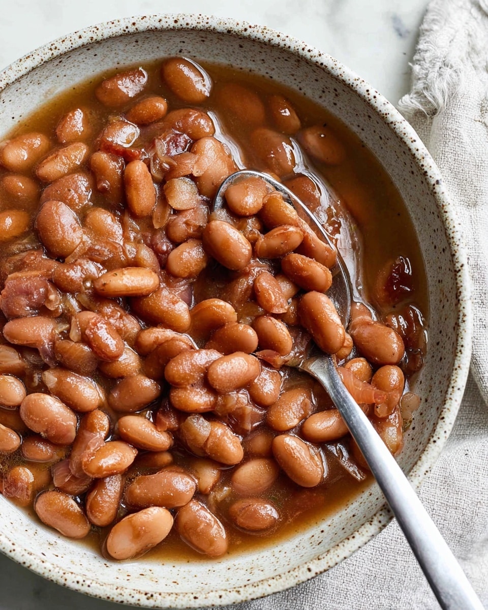 A close-up image of a white speckled bowl filled with cooked pinto beans in a brownish, glossy sauce. The beans are soft and varied in size with some skin slightly peeling. A silver spoon rests inside the bowl, partially covered by the beans and sauce. The bowl sits on a white marbled surface with a light-colored cloth partially visible in the top right corner. photo taken with an iphone --ar 4:5 --v 7