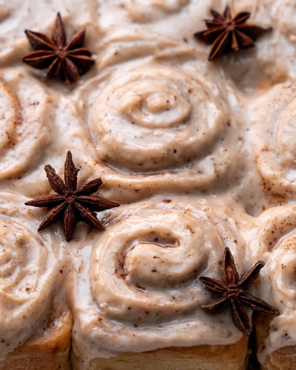 The image shows a close-up of a group of cinnamon rolls covered in a thick, creamy frosting with a light brown color and specks of cinnamon throughout. There are visible swirls in the rolls beneath the smooth, shiny frosting that looks soft and slightly glossy. Two star anise spices are placed on top as decoration, adding a dark brown contrast to the light frosting. The rolls are packed closely, creating visible round shapes from the spiraled dough underneath the frosting. The background is a white marbled texture. photo taken with an iphone --ar 4:5 --v 7