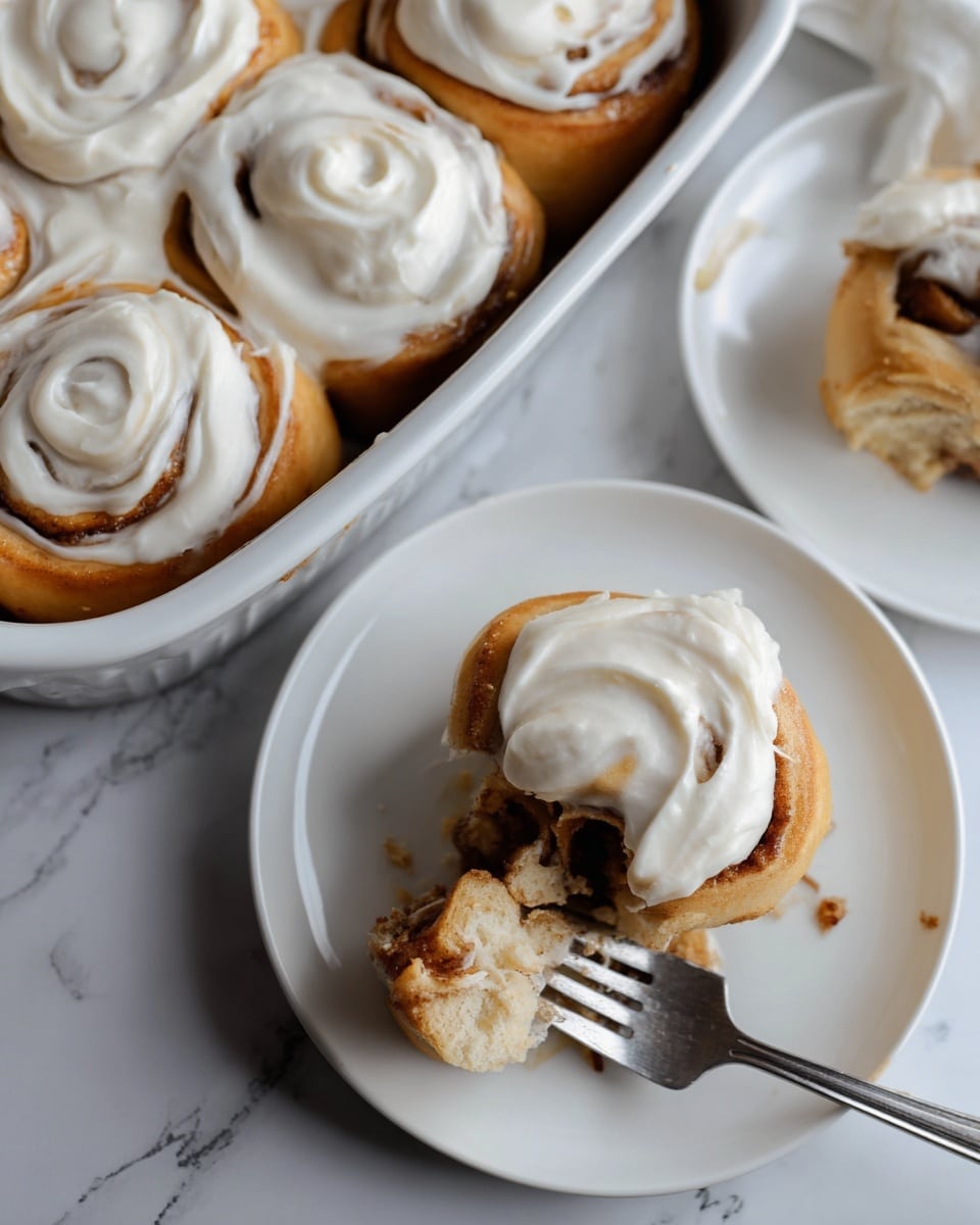 The image shows soft cinnamon rolls with a thick layer of creamy white frosting swirled on top of each roll, highlighting the spiral shape and light brown dough underneath. The rolls are placed on white round plates and in a white oval baking dish, all set on a white marbled surface. One cinnamon roll is partially eaten with a silver fork holding a bite-sized piece, showing the fluffy texture and cinnamon filling. The overall look is warm and inviting with smooth frosting contrasting the slightly textured dough. Photo taken with an iphone --ar 4:5 --v 7