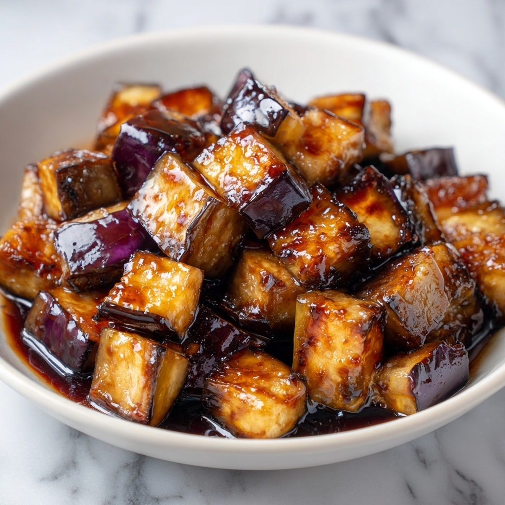 A close-up view of a dish with many small, shiny cubes of cooked eggplant on a white plate. Each cube shows dark purple skin on some sides and a golden-brown, slightly crispy and glazed surface on others, covered with a thick, dark sauce that looks sticky and rich. The cubes are piled high in the center of the plate, with the sauce glistening in the light, creating a glossy texture. The background is a white marbled texture. photo taken with an iphone --ar 4:5 --v 7