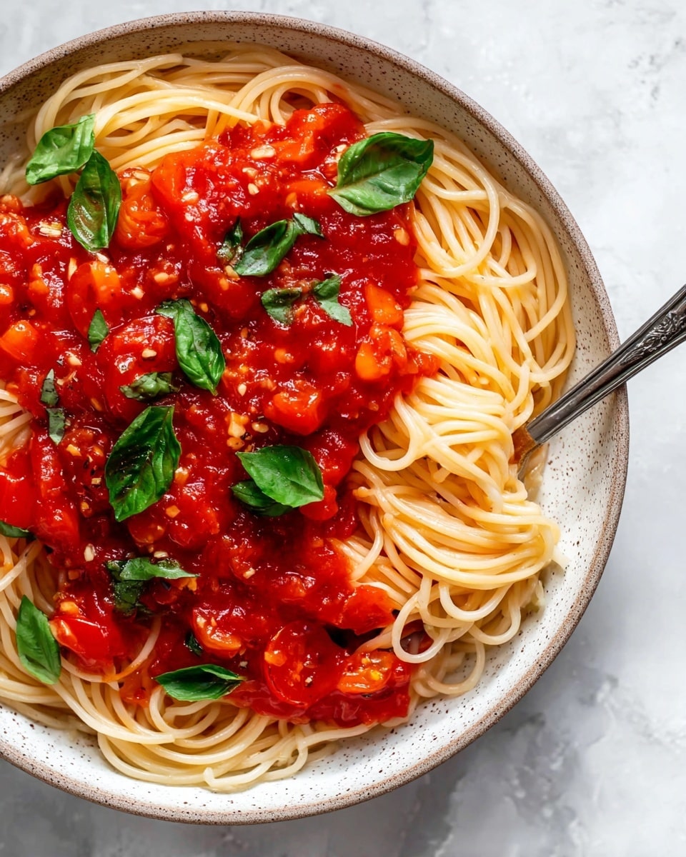 A white speckled bowl filled with thin, light yellow spaghetti noodles forming the base layer, topped with a bright red chunky tomato sauce containing pieces of cooked tomatoes and small bits of garlic. Fresh green basil leaves are scattered on top, adding a fresh contrast to the red sauce and light noodles. A silver fork rests inside the bowl, partially hidden by the noodles and sauce, all placed on a white marbled surface. photo taken with an iphone --ar 4:5 --v 7