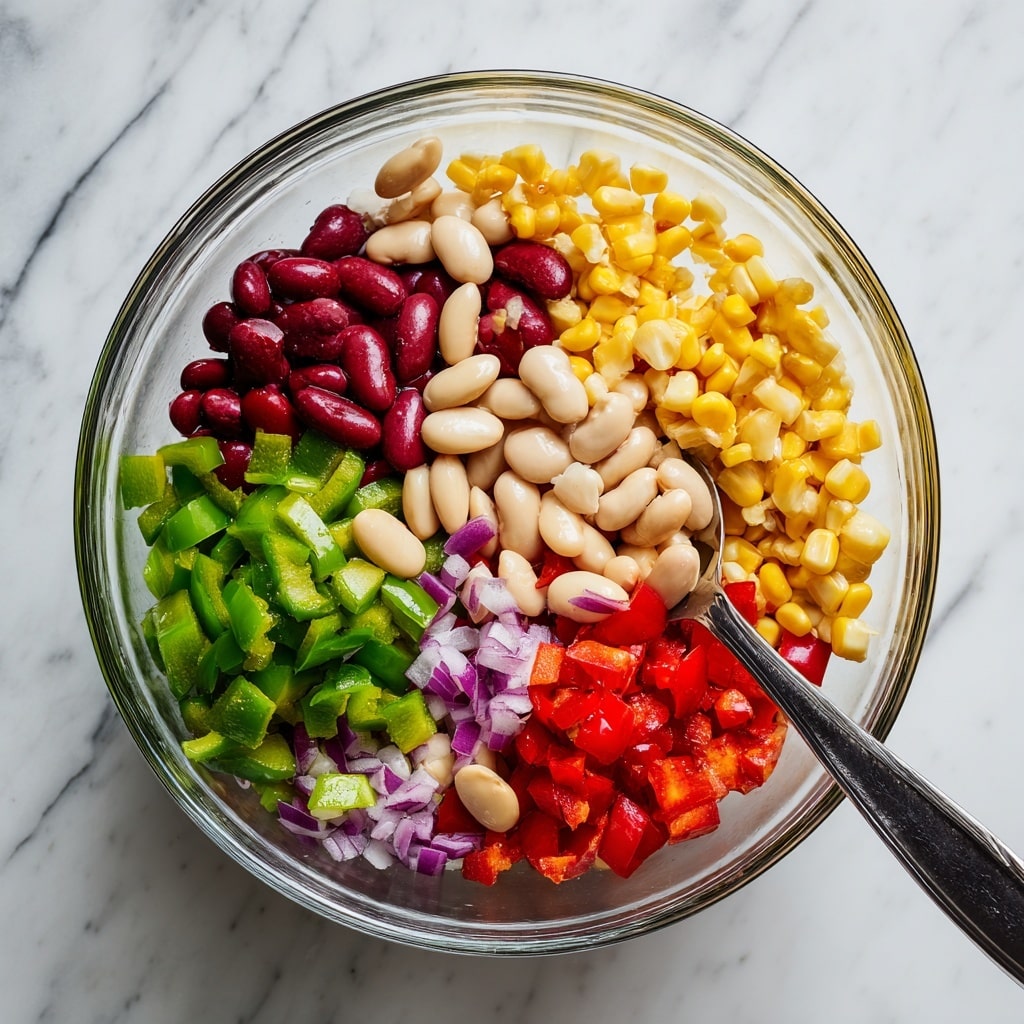 A glass bowl sits on a white marbled surface, filled with a colorful mix of salad ingredients. The salad has several layers visible from the top: red kidney beans and white beans with smooth and glossy textures, scattered among bright yellow corn kernels that look soft and plump, chopped green bell peppers with a fresh, slightly shiny green surface, small pieces of red bell pepper adding vibrant color, and finely diced purple onions that add a bit of sharpness with their slightly translucent quality. A silver spoon with a dark handle rests inside the bowl on the left side. photo taken with an iphone --ar 4:5 --v 7