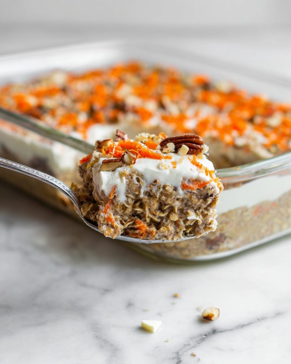 A close-up view shows a spoon holding a layered oatmeal dish with a thick base layer that looks soft and grainy, filled with brown oats and bits of pecans. On top of this base is a creamy white layer, likely yogurt or cream, sprinkled with bright orange shredded carrot bits and small chunks of pecan nuts, adding texture and color. Behind the spoon is a clear rectangular glass baking dish filled with the same layered oatmeal mix, cream, and carrot pieces, sitting on a white marbled surface. Small white pieces are scattered near the spoon in the foreground. The photo taken with an iphone --ar 4:5 --v 7