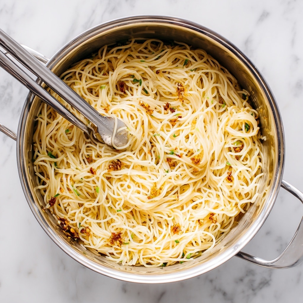A pot filled with thin, light yellow noodles that look soft and slightly shiny, mixed with small bits of crispy brown garlic and tiny green pieces, probably scallions. The noodles are tangled and loosely packed, covering the whole inside of the pot. Two silver tongs rest inside, one partially buried in the noodles, and the pot sits on a white marbled surface. photo taken with an iphone --ar 4:5 --v 7