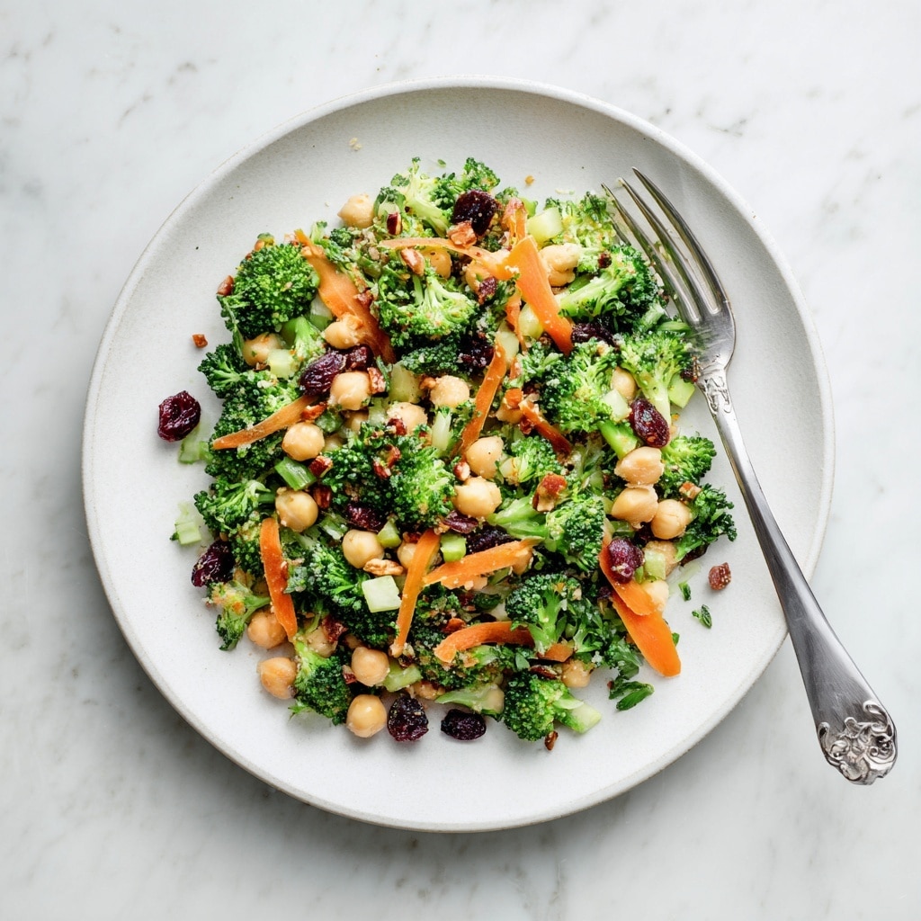A fresh salad is served on a white plate, placed on a white marbled surface. The salad has several colorful layers including small broccoli florets in green, thin carrot strips in bright orange, chickpeas in light tan, dark dried berries, and small pieces of nuts with a brown shade. The vegetables and toppings are mixed evenly, showing textures from crunchy broccoli and nuts to the smooth chickpeas. A metal fork rests on the right side of the plate, partially touching the food. Small bits of herbs and salad ingredients are scattered lightly around the plate edges. photo taken with an iphone --ar 4:5 --v 7