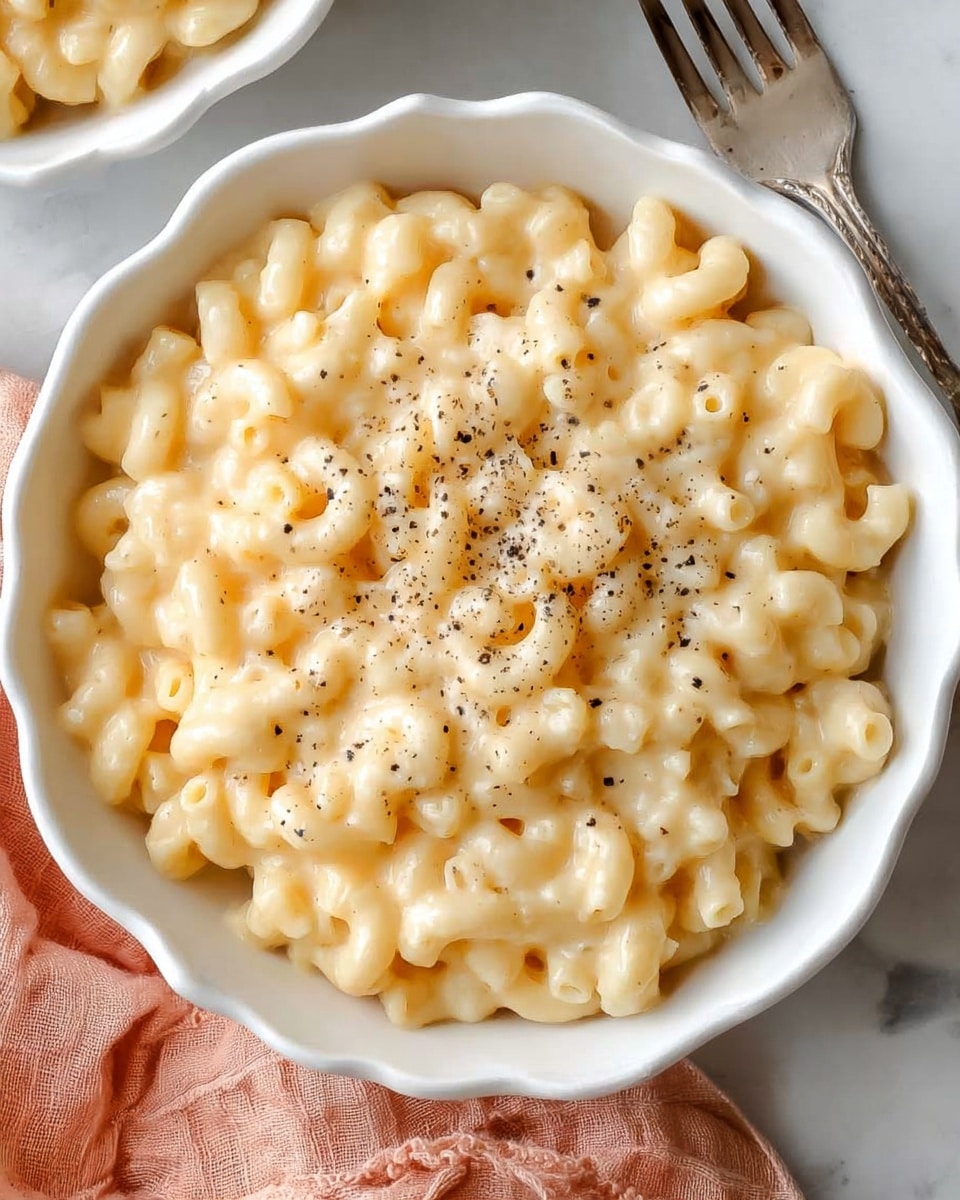 A close-up view of two white bowls filled with creamy macaroni and cheese, each bowl containing soft, elbow-shaped pasta coated in a smooth, pale yellow cheese sauce sprinkled with small specks of black pepper; one bowl is in the front, slightly to the right, with a fork lifting a forkful of macaroni, held by a woman's hand from the right side, while the second bowl is in the back, slightly blurred; the setup is on a white marbled surface with a light brown wooden pepper grinder behind the bowls and a folded dusty pink cloth napkin in the lower left corner. photo taken with an iphone --ar 4:5 --v 7