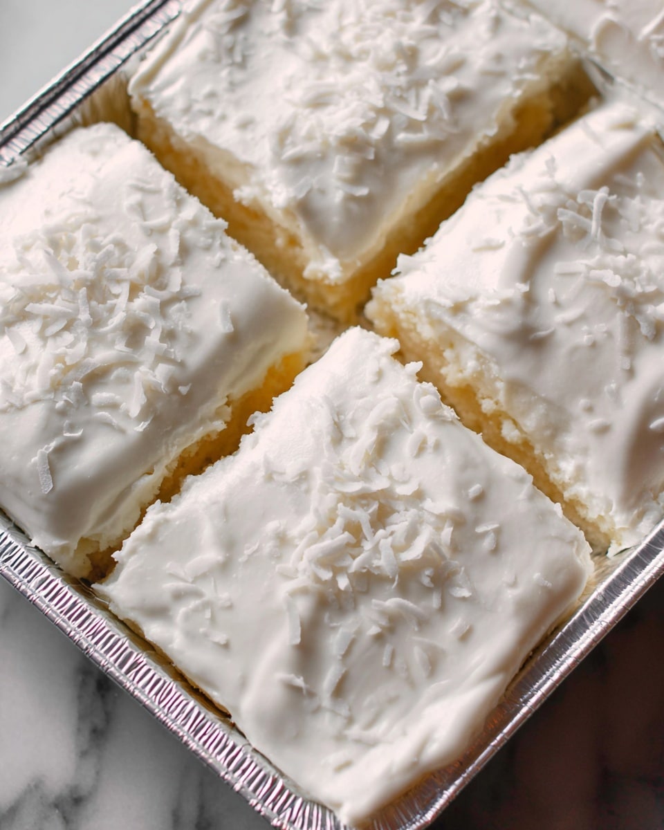 The image shows four square pieces of a dessert in a silver foil pan, each topped with a thick, smooth layer of white frosting. The frosting is spread evenly over the top, with a soft and creamy texture. Small, thin coconut shreds are scattered lightly across the frosting, adding a delicate texture and extra layer of white on each piece. The dessert pieces are closely set together, with clear, sharp edges dividing them. The background is a white marbled surface. Photo taken with an iphone --ar 4:5 --v 7