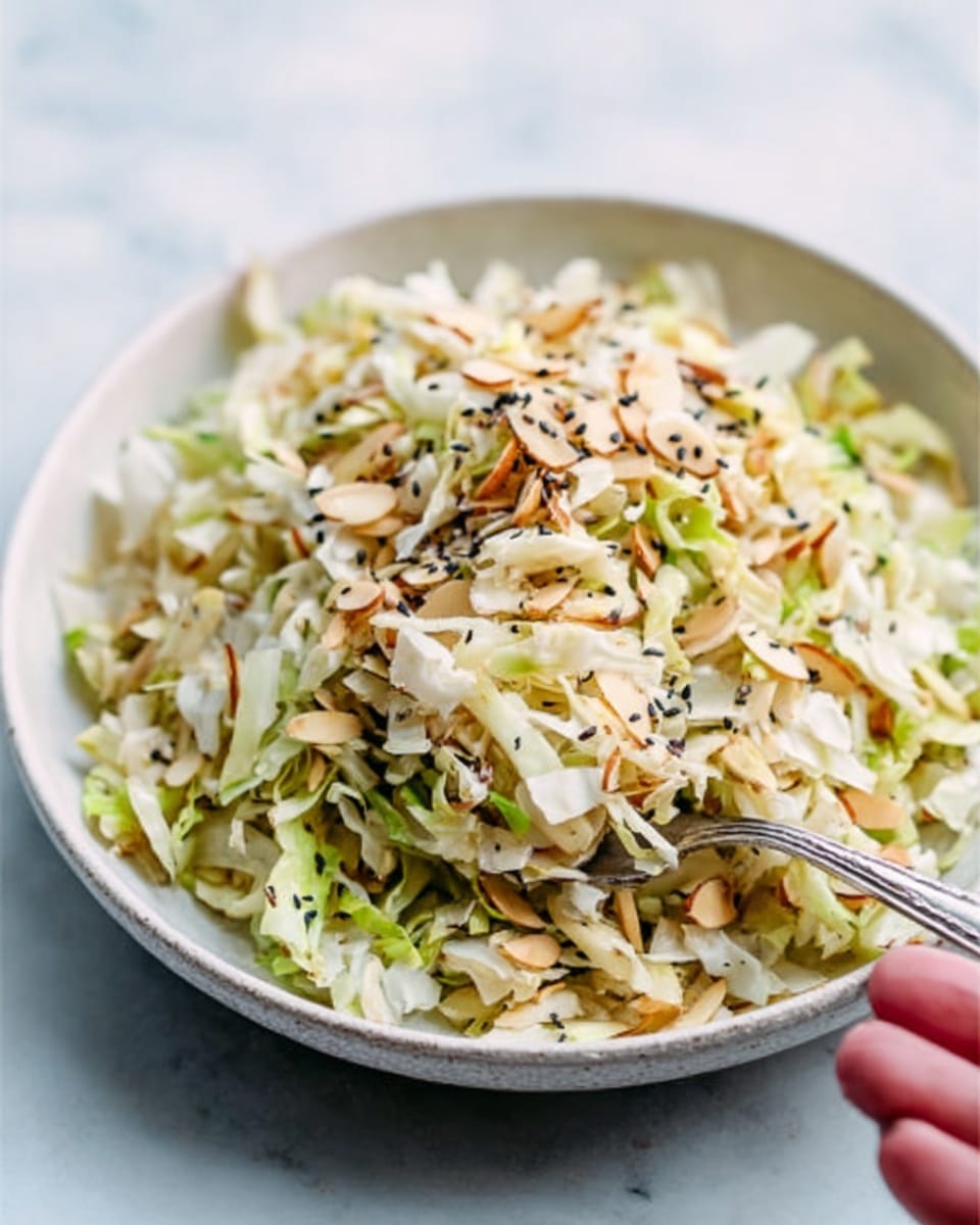 A close-up of a white bowl filled with a mixture of shredded pale cabbage and light beige slivered almonds, topped with some black sesame seeds scattered evenly across the surface. A silver fork rests inside the bowl, with a woman's hand holding it gently from the side. The bowl sits on a white marbled surface that brightens the whole scene. The colors are soft and natural, with light textures from the crunchy almonds and shredded cabbage. Photo taken with an iphone --ar 4:5 --v 7