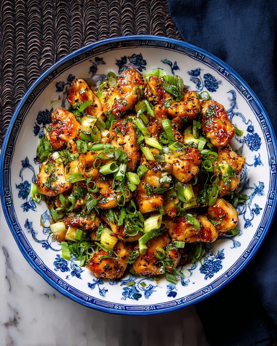 A white plate with blue floral design holds a dish of cooked chicken pieces, browned and glistening with a shiny, oily sauce. The chicken is mixed with chopped bright green onions scattered on top and throughout. The chicken pieces look tender with slightly crisp edges, and the sauce has a rich reddish-brown color mixed with green from the onions, giving the dish a lively and flavorful appearance. The plate is set on a white marbled textured surface. photo taken with an iphone --ar 4:5 --v 7