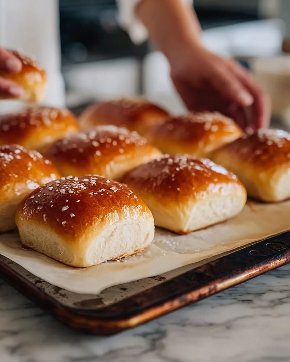 The image shows nine golden-brown bread rolls with a shiny, slightly crisp top sprinkled with small white granules, likely sugar or salt. The rolls are square-shaped with a soft, fluffy white inside and are placed on a parchment-lined white baking tray that has a dark, worn edge. The tray is resting on a surface with a white marbled texture. In the background, a woman's hands are visible, reaching towards the tray as if about to pick up one of the rolls. photo taken with an iphone --ar 4:5 --v 7