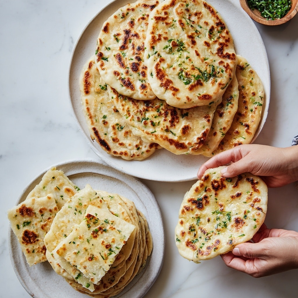 The image shows two sets of garlic flatbreads on white plates placed on a white marbled surface. The top set consists of many round, lightly browned flatbreads with green herbs sprinkled on top, stacked slightly overlapping in a row. A woman's hand is holding one flatbread at the right side. The flatbreads have a soft, slightly puffy texture with patches of golden brown. The bottom set shows three stacks of flatbreads: two stacks of whole flatbreads and one stack of flatbreads cut into square pieces. A woman's hand is holding a torn flatbread above the cut pieces, showing its soft and fluffy inside with green herb spots. Below these images, there is text with a beige paper-like background listing ingredients for making the flatbreads. Photo taken with an iphone --ar 4:5 --v 7
