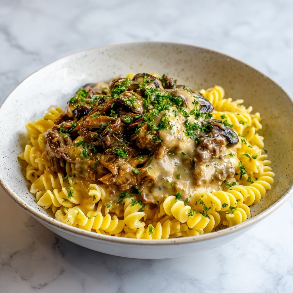A close-up of a white bowl filled with two main layers: the bottom layer has light yellow egg noodles with a slightly twisted shape and a soft texture, scattered with green chopped herbs; the top layer is a rich brown mixture of shredded beef and sliced mushrooms covered in a glossy, creamy sauce, also sprinkled with chopped green herbs. The bowl rests on a white marbled surface. photo taken with an iphone --ar 4:5 --v 7