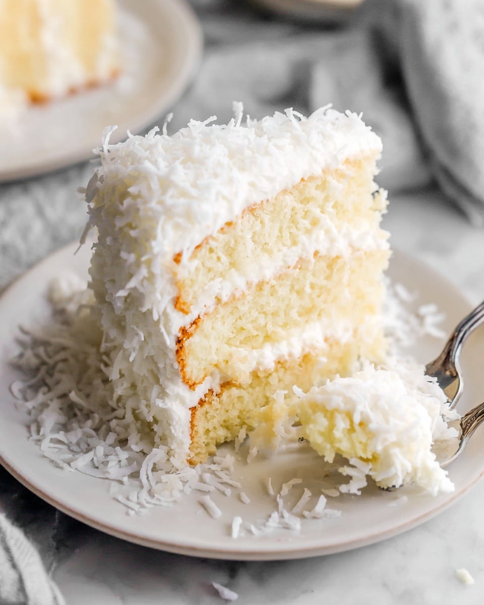 A slice of white layered cake sits on a white plate scattered with white coconut flakes. The cake has three visible layers: the top and bottom are soft, light yellow sponge cake, while the middle is a creamy yellow filling. The outside is covered in thick white frosting with a fluffy texture, topped and surrounded by shredded coconut. A fork is cutting into the cake from the lower left side. The background is a white marbled texture with another blurred white plate and cloth visible. Photo taken with an iphone --ar 4:5 --v 7