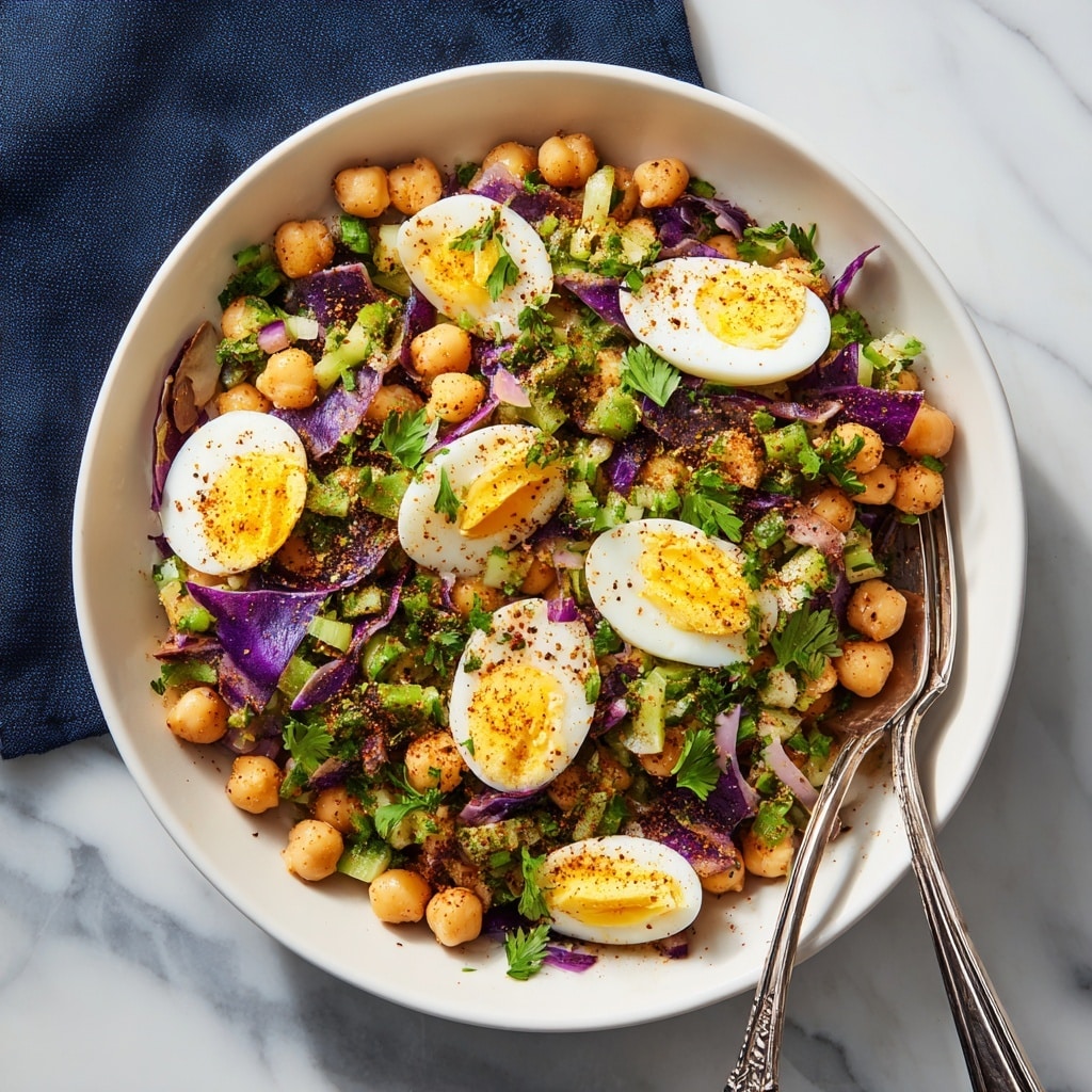A white bowl holds a chickpea salad made of a bottom layer of round, light beige chickpeas, mixed with chopped green celery and purple cabbage pieces spread evenly. On top are slices and rings of hard-boiled eggs with smooth white edges and bright yellow yolks scattered across. Bright green parsley leaves are sprinkled generously, adding freshness. The dish is sprinkled with a dark spice powder for contrast. Two vintage silver forks rest on the left rim of the bowl, and the bowl sits on a white marbled surface with a dark blue cloth partially visible on the upper left side. Photo taken with an iphone --ar 4:5 --v 7