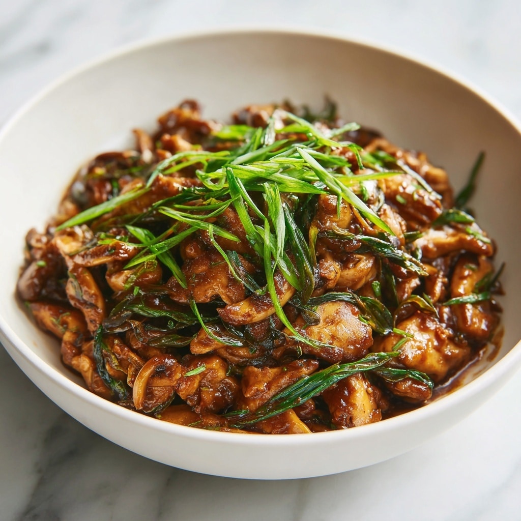 A close-up image of a white bowl filled with a layered dish made mainly of small pieces of cooked chicken mixed with thin, long brown mushrooms in a thick, glossy brown sauce. The dish is topped with bright green chopped spring onions, giving a fresh color contrast. The bowl sits on a white marbled surface, and soft natural light highlights the glossy texture of the sauce and freshness of the green onions. Photo taken with an iphone --ar 4:5 --v 7