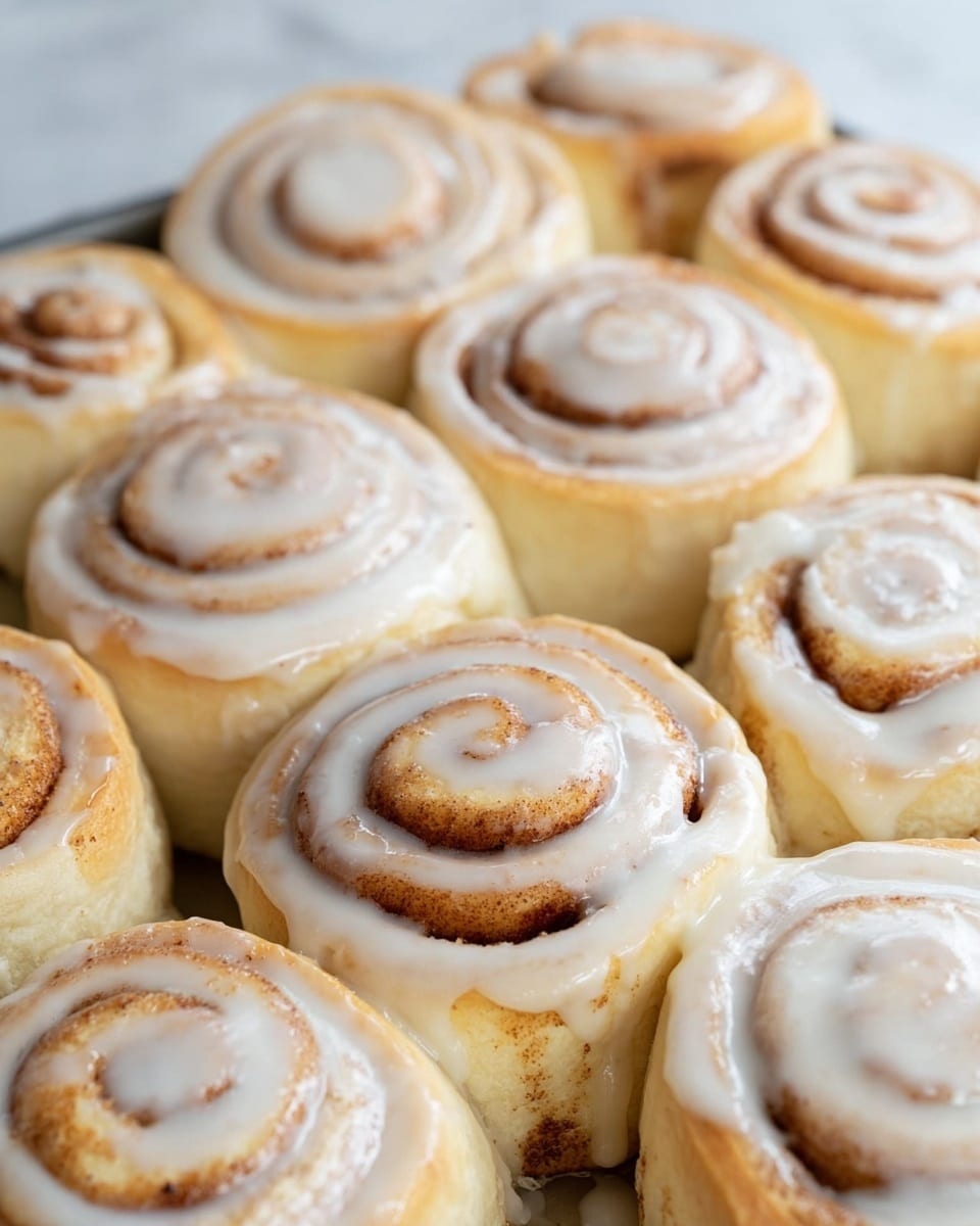 The image shows a close-up of nine cinnamon rolls placed closely together in a baking pan. Each roll has visible swirled layers with a light golden brown color mixed with white creamy icing that follows the spiral pattern. The rolls are soft and fluffy, with some spots showing a slightly darker cinnamon filling. The icing is smooth and glossy, evenly spread over each roll’s top, with some drips down the sides. The background surface is a white marbled texture. photo taken with an iphone --ar 4:5 --v 7