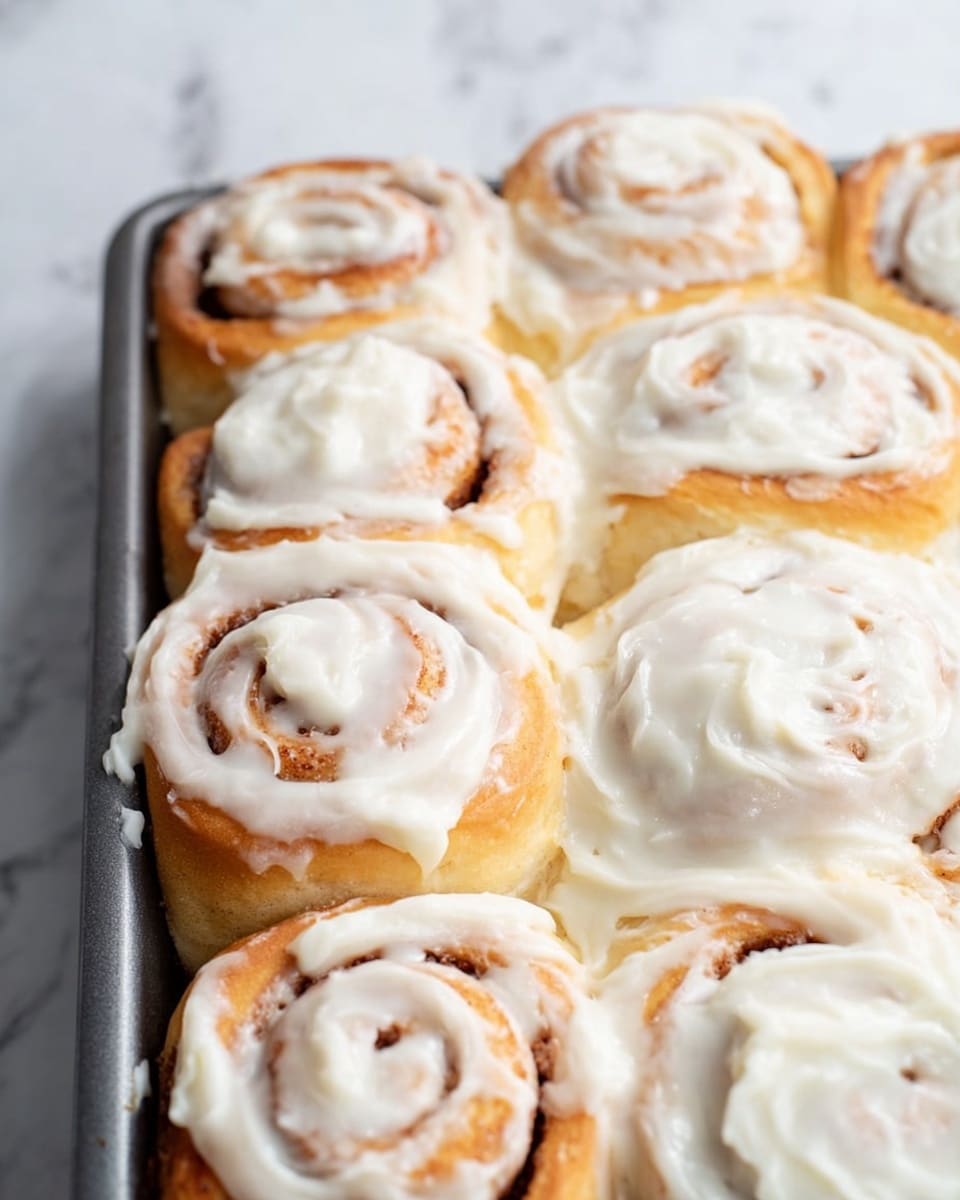 A close-up view of a pan filled with cinnamon rolls, each roll having a golden-brown spiral base layer with visible swirls of cinnamon filling. On top of the cinnamon base, a thick layer of white cream cheese frosting generously covers most of the rolls, dripping slightly around the edges and filling the gaps between them. The pan is placed on a white marbled surface which adds a clean and bright background to the image. Photo taken with an iphone --ar 4:5 --v 7