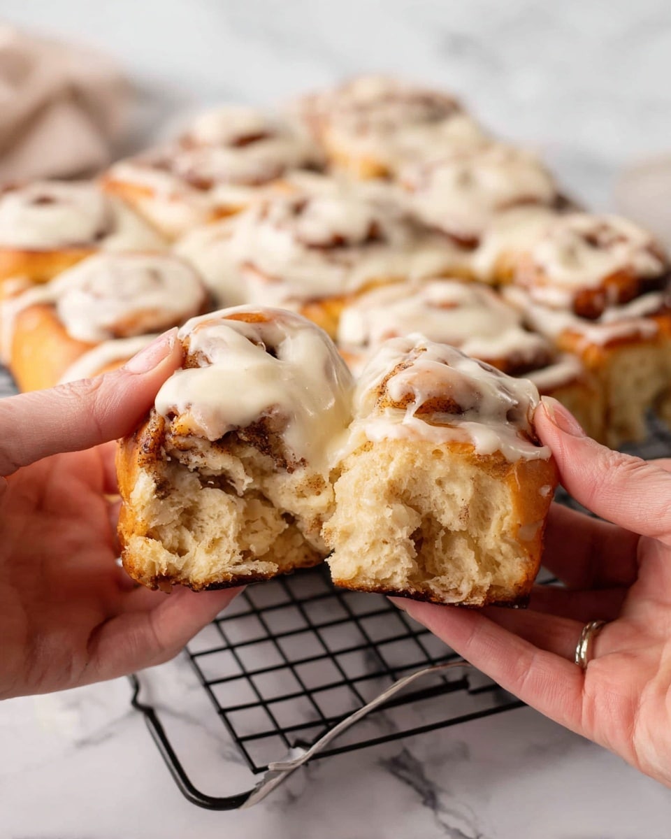 A close-up of a pair of woman's hands pulling apart a soft, thick cinnamon roll covered in creamy white icing. The cinnamon roll has an uneven texture with layers of golden-brown dough swirled with darker cinnamon filling. In the background, a white icing-covered batch of multiple cinnamon rolls is placed together on a black cooling rack over a white marbled surface. The contrast between the warm brown cinnamon spots and the smooth, shiny white icing is clear. Photo taken with an iphone --ar 4:5 --v 7