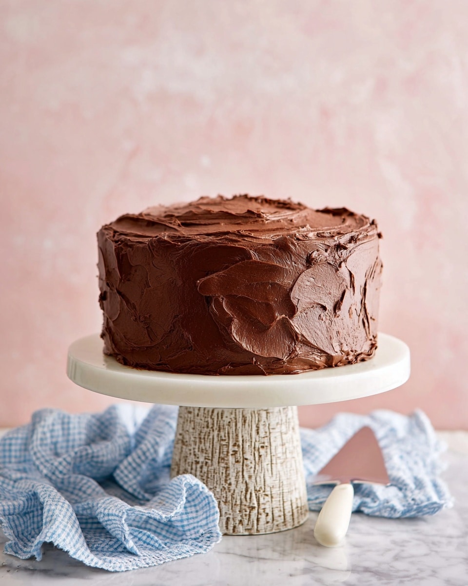 The image shows a round chocolate cake with thick, rich, smooth chocolate frosting spread unevenly around the sides and top, creating a textured look with swirls and ridges. The cake has at least two layers inside, hinted by its height and structure. It sits on a white cake stand with a tree trunk-like textured base, placed on a white marbled surface. Next to the stand is a blue and white checkered cloth under a white ceramic cake server. The background is a soft pale pink color. photo taken with an iphone --ar 4:5 --v 7