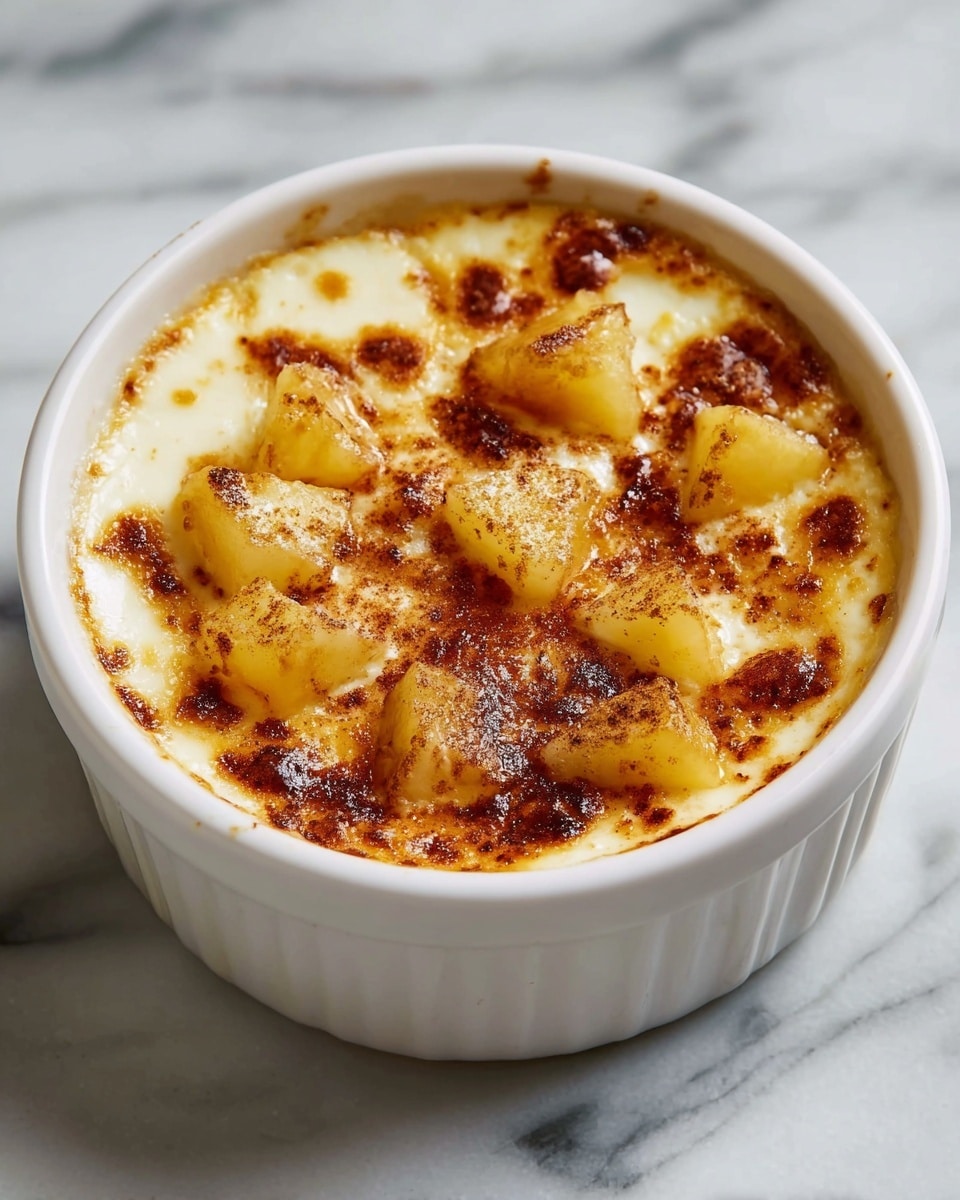 A white ramekin filled with a creamy, baked dessert that has a golden-brown, lightly caramelized top layer with some darker spots. Scattered on top are chunks of soft, cooked yellow fruit pieces with a dusting of brown cinnamon powder, adding texture and warmth. The dessert looks smooth and slightly bubbly around the edges, sitting on a white marbled surface. photo taken with an iphone --ar 4:5 --v 7