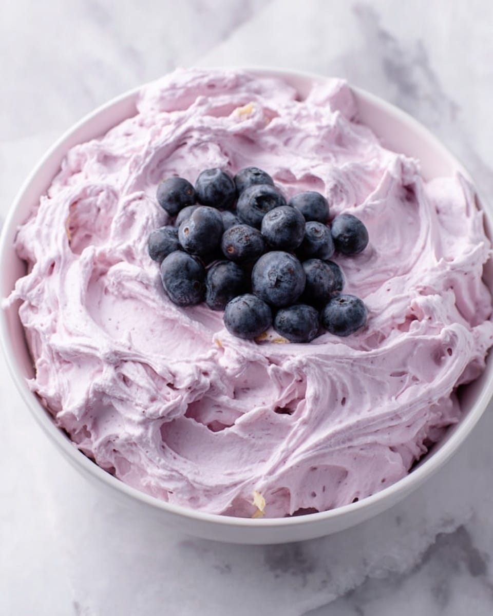 A white bowl filled with a creamy, light purple mixture that looks soft and thick. The top layer is decorated with fresh, round blueberries evenly spread out and small, white marshmallows scattered among the berries. A metal spoon is placed inside the bowl, slightly sinking into the mixture. The bowl sits on a light-colored, textured surface that looks clean and smooth. In the background, another similar bowl is partially visible, filled with the same mixture and toppings. Photo taken with an iphone --ar 4:5 --v 7