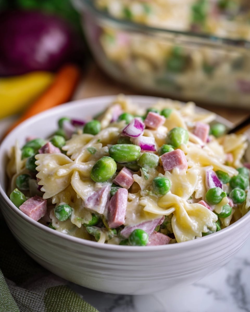 This image shows a close-up of a creamy pasta salad served in a white bowl. The dish has several layers: on the bottom, tender bowtie pasta with a pale yellow color, mixed with bright green peas scattered throughout. Thin slices of purple-red onion and small chunks of pink ham are mixed evenly with the pasta and peas. Fresh green snow peas and thin slices of white and red radish add texture and color contrast. The creamy dressing coats all the ingredients, giving them a shiny, smooth texture. The bowl sits on a white marbled surface, with part of another bowl of the same salad visible at the bottom. A silver fork rests near the upper bowl on the surface. Photo taken with an iphone --ar 4:5 --v 7