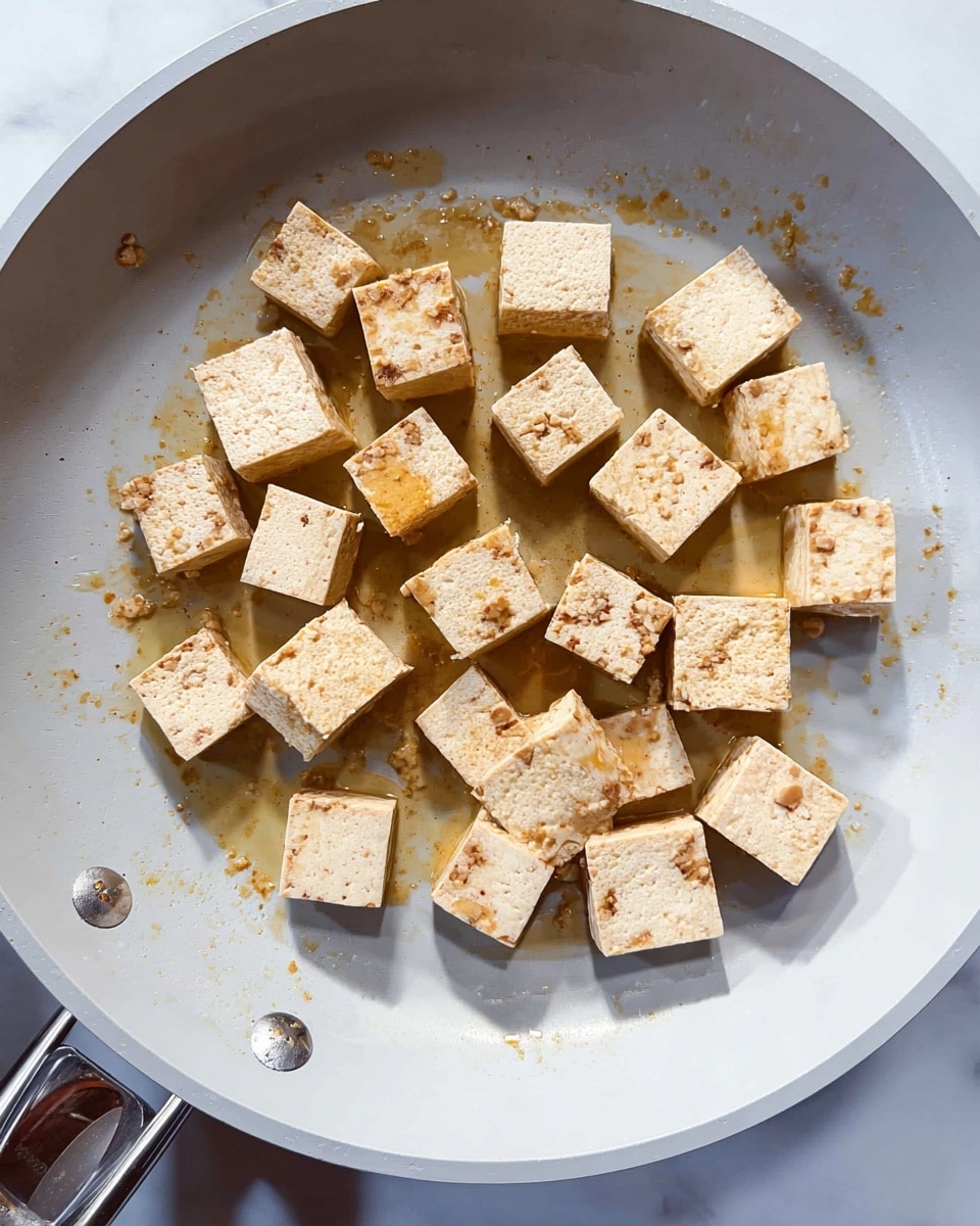 The image shows small, light beige tofu cubes evenly spaced in a white frying pan, some with a slight golden brown sear on one side, indicating they are being cooked. The cubes have a soft and smooth texture with tiny indentations. A little golden oil glistens around the tofu pieces, contrasting with the light grey surface of the pan. The pan handle is visible in the lower left corner, and the scene sits on a white marbled surface. photo taken with an iphone --ar 4:5 --v 7