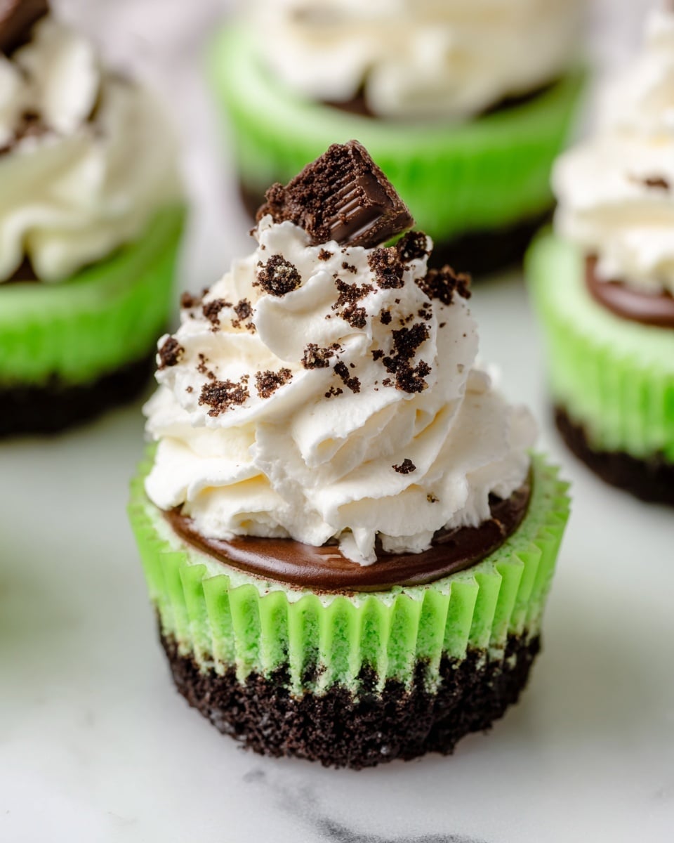 The image shows a stack of three mini cheesecakes on a wooden board with a white marbled background. Each cheesecake has two layers: a dark brown crumbly crust at the bottom and a thick, light green creamy layer on top, speckled with darker bits. The top cheesecake is decorated with a swirl of white whipped cream sprinkled with small dark crumbs and topped with a piece of dark chocolate. The middle cheesecake has a bite taken out, showing the creamy green filling inside. Small chocolate chips and green wrapped chocolates lie beside the stack. Photo taken with an iphone --ar 4:5 --v 7