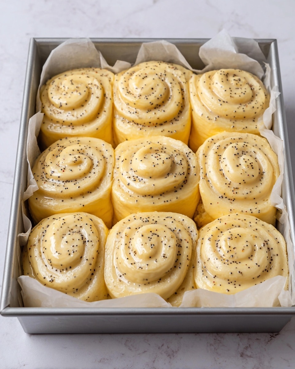 The image shows a silver baking tray lined with white parchment paper, filled with twelve uncooked swirl dough rolls arranged in three rows. Each roll is smooth and glossy with a pale yellow color, speckled evenly with small black poppy seeds throughout the spiral layers. The dough looks soft and slightly sticky, with the swirls forming three to four visible layers from top to bottom. The tray is placed on a white marbled surface, creating a clean and bright background. photo taken with an iphone --ar 4:5 --v 7