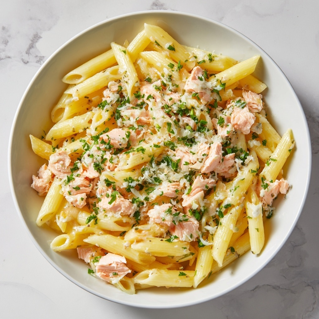 A white bowl filled with three main layers is shown on a white marbled texture. The bottom layer is pale yellow penne pasta with a smooth, slightly glossy texture. The middle layer has small pieces of light pink cooked salmon, scattered evenly among the pasta. The top layer is sprinkled with finely grated white cheese and small bright green parsley bits, adding contrast and freshness to the dish. Photo taken with an iphone --ar 4:5 --v 7