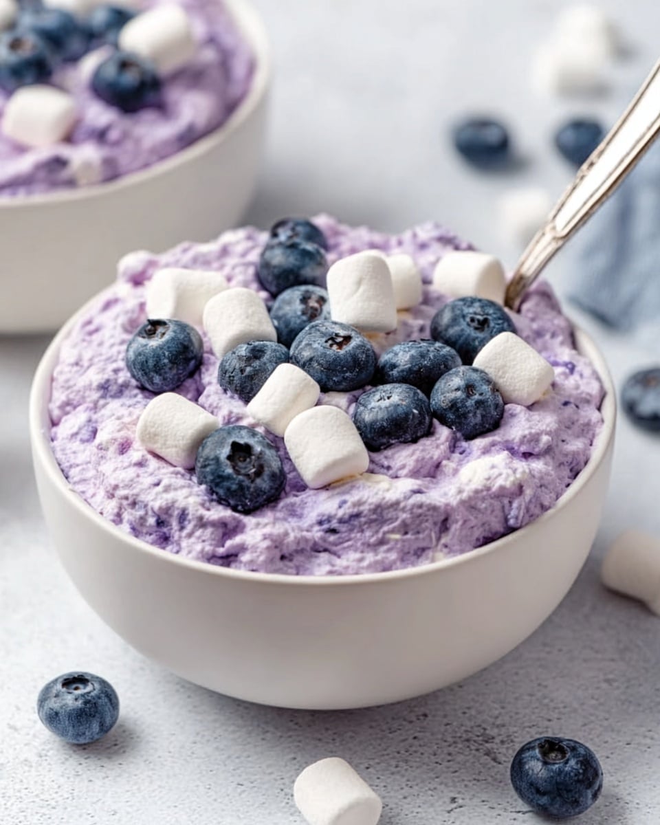 A white bowl filled with thick, creamy light purple mousse that has a soft, whipped texture with swirled peaks all over the surface. In the center, there is a small pile of fresh, round, dark blue blueberries that add contrast and a pop of color. The bowl is placed on a white marbled surface. Photo taken with an iphone --ar 4:5 --v 7