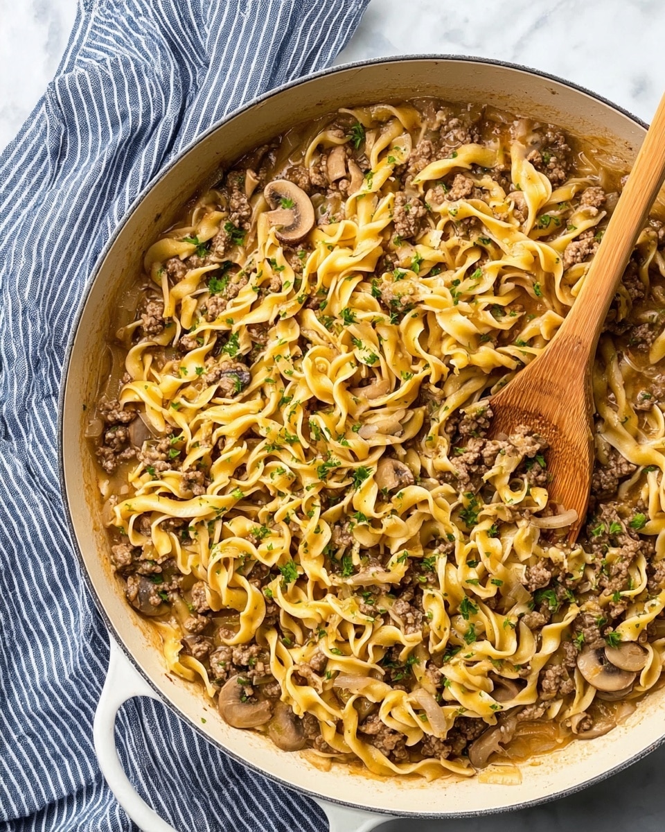A large white pan filled with cooked egg noodles mixed with ground beef and thin slices of mushrooms, all coated in a light brown sauce. The noodles are twisted and soft, scattered evenly throughout the pan with small bits of cooked onions visible. Fresh green parsley is sprinkled over the top, adding a touch of color. A wooden spoon rests inside the pan, partially covered by the noodles. The pan is placed on a white marbled surface with a blue and white striped cloth next to it. Photo taken with an iphone --ar 4:5 --v 7