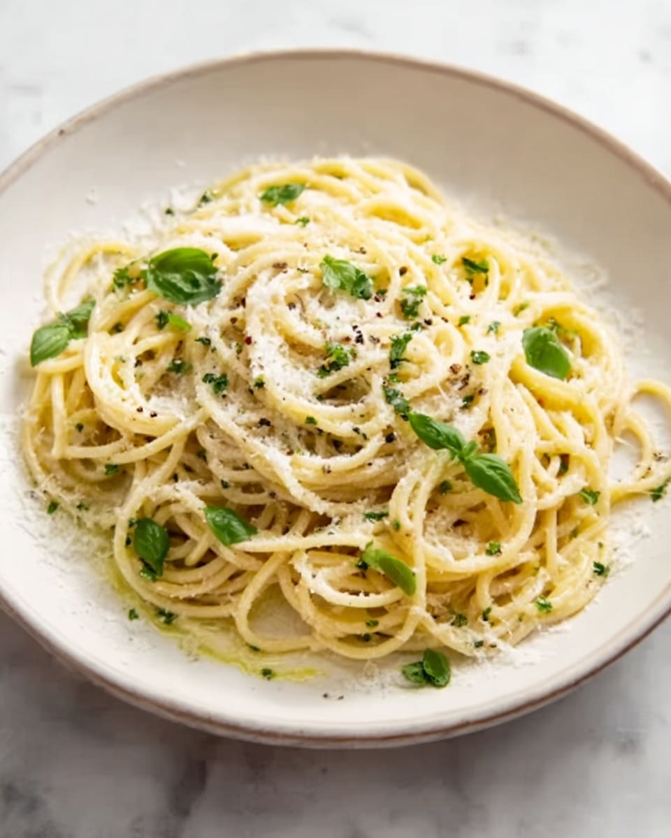 A white bowl holds a serving of spaghetti coated in a creamy white sauce, with thin lemon peel curls scattered on top. Fresh bright green basil leaves are placed both on the pasta and around the bowl, adding vibrant color. Black pepper is lightly sprinkled over the dish. The bowl sits on a white marbled texture with a wooden cutting board nearby holding a piece of cheese and a grater. A lemon wedge and a small bowl of grated cheese rest beside a fork with a black handle, completing the scene. Photo taken with an iphone --ar 4:5 --v 7