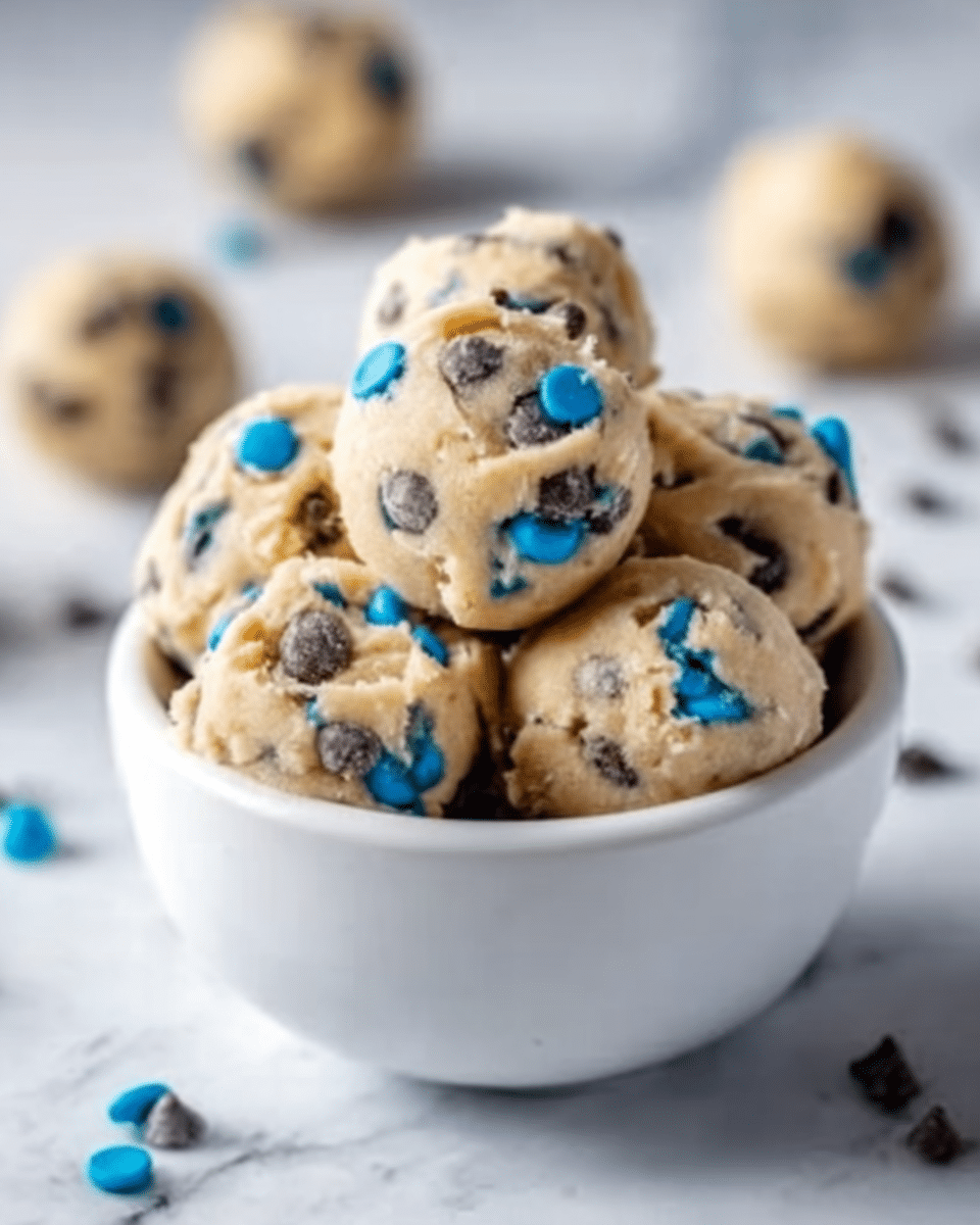 A white bowl filled with several round cookie dough balls, each studded with chocolate chips and colorful blue candy pieces. The dough balls are soft and slightly textured, piled neatly inside the bowl. The background shows a white marbled surface with a few scattered cookie dough balls blurred in the distance. Photo taken with an iphone --ar 4:5 --v 7