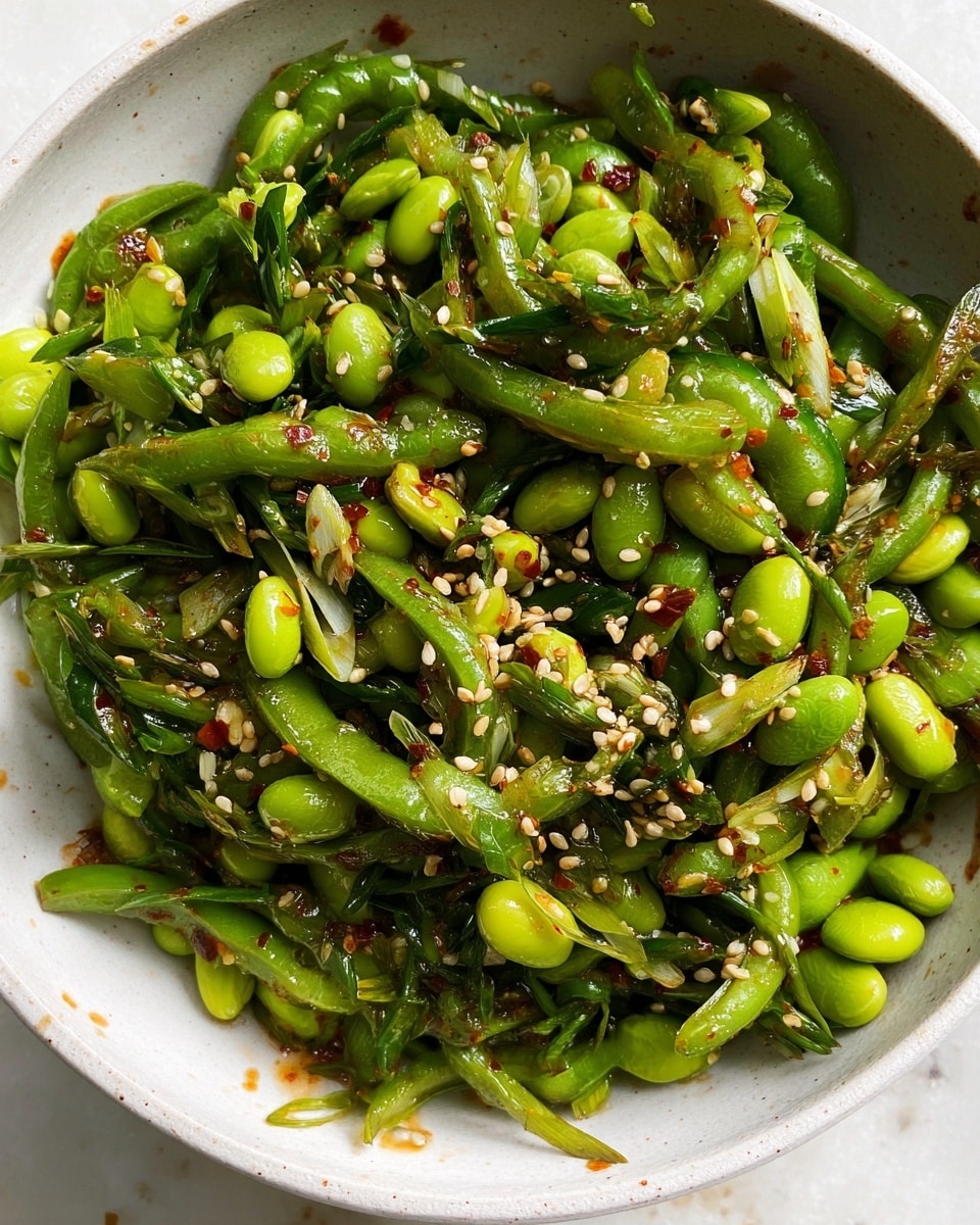 A close-up view of a white bowl filled with a vibrant green salad made mainly of sliced green beans, edamame beans, and chopped green onions. The salad is tossed with sesame seeds and small bits of red chili flakes, adding specks of white and red throughout. The beans and onions have a fresh, glossy texture, suggesting a light dressing. The bowl rests on a white marbled surface with a few scattered green and red bits around the edge of the bowl. photo taken with an iphone --ar 4:5 --v 7
