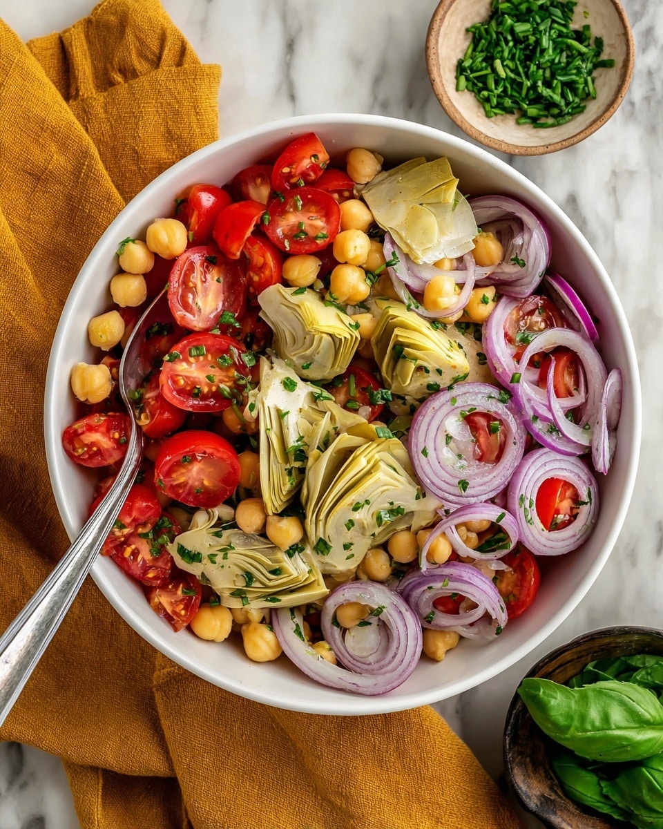 A white bowl filled with a colorful salad featuring four main layers: the bottom layer is small, pale yellow chickpeas, topped with bright red halved cherry tomatoes, thin rings of translucent purple-red onion, and quartered pale yellow artichoke hearts with a soft texture. Scattered small green chives add specks of fresh green color throughout. A silver spoon is placed inside the bowl. The bowl sits on a white marbled surface with a mustard-colored cloth beside it, a small white bowl of chopped chives above, and a dark bowl of fresh green basil leaves below. Photo taken with an iphone --ar 4:5 --v 7