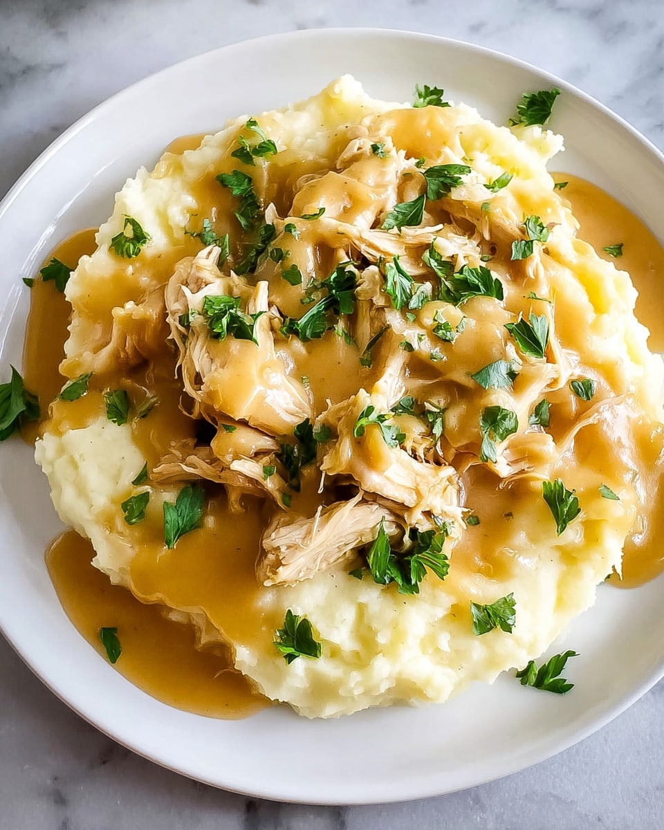 A white plate holds a dish with two main layers; the bottom layer is a bed of soft, pale yellow mashed potatoes with a slightly lumpy texture, and the top layer consists of shredded chicken coated in a thick, creamy brown gravy. Bright green parsley pieces are scattered on top, adding a fresh contrast of color. The plate sits on a white marbled textured surface with part of a green-and-white striped cloth visible in the background. Photo taken with an iphone --ar 4:5 --v 7