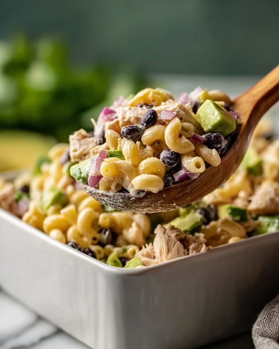 A close-up view of a bowl filled with creamy pasta salad, featuring three main layers: the top layer has light green avocado chunks with a soft texture, the middle layer shows black olives and bright yellow corn kernels scattered throughout, and the base layer consists of beige cavatappi pasta pieces coated with a creamy sauce, mixed with small bits of chopped red onion and green herbs. The bowl is white with a natural pottery look, sitting on a white marbled surface with a piece of beige linen fabric beside it. Photo taken with an iphone --ar 4:5 --v 7