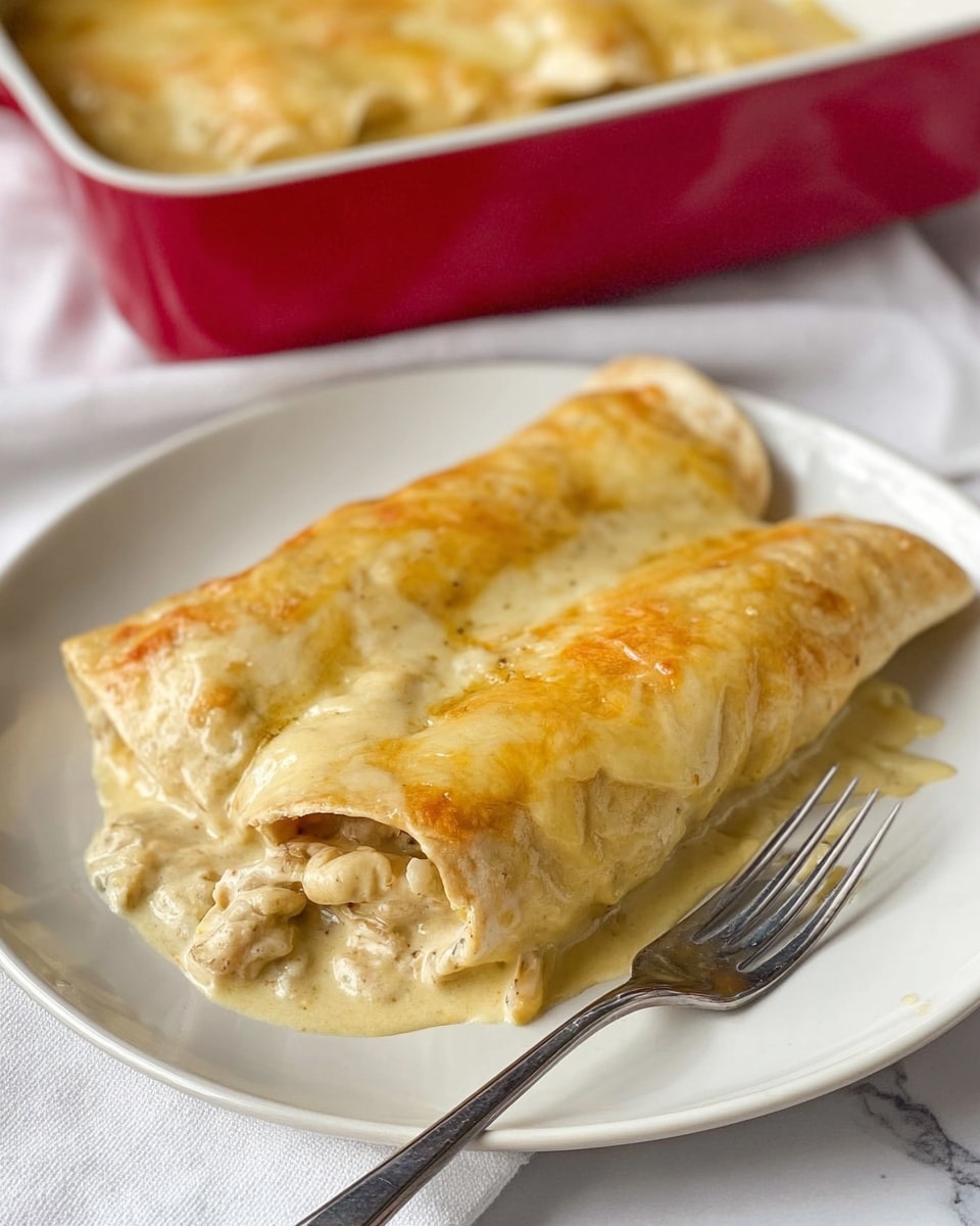 Two enchiladas are served on a plain white plate, covered in a creamy, light yellow sauce with a smooth, slightly bubbly texture. The enchiladas have a soft, folded tortilla layer visible beneath the sauce, with the front one partially open to show a creamy filling mixed with small chunks, all that is pale in color. A silver fork is placed under the open enchilada on the right side of the plate. The plate sits on a white cloth with a white marbled surface in the background, and a red baking dish holding more enchiladas is visible but out of focus behind the plate. Photo taken with an iphone --ar 4:5 --v 7