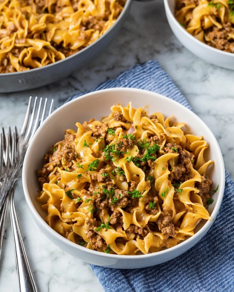 A white bowl filled with three layers of cooked egg noodles mixed with ground beef and small diced onions, all coated in a light brown sauce. The noodles are pale yellow with a soft texture, curled and layered evenly with browned ground beef pieces. The dish is topped with a sprinkling of fresh chopped green parsley for contrast. The bowl sits on a white marbled surface next to a blue striped cloth napkin, with two silver forks nearby. In the background, there is a white bowl and a gray pan partially visible, both containing the same noodle dish. Photo taken with an iphone --ar 4:5 --v 7