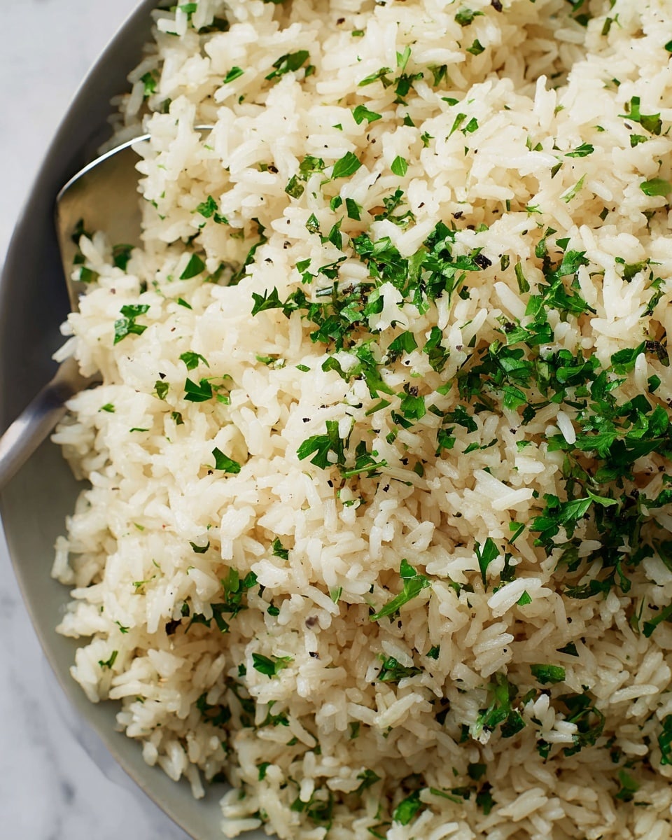 The image shows a close-up of cooked rice mixed with finely chopped green herbs evenly scattered throughout. The rice grains are white to slightly off-white, soft, and fluffy with a light, almost translucent texture. The green herbs, possibly parsley or cilantro, add a fresh pop of color and small black pepper specks are visible, adding texture contrast. The rice mixture appears moist but not wet, and a silver spoon is partially visible, gently scooping some of the rice. The scene has a bright and clean appearance with the dish set against a white marbled surface. photo taken with an iphone --ar 4:5 --v 7