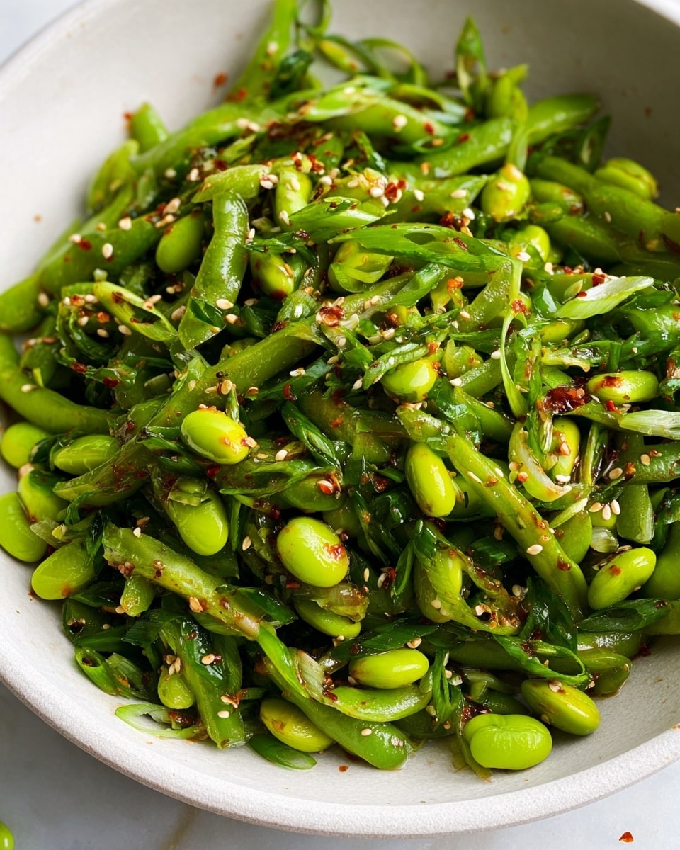 The image shows a close-up of a white bowl filled with a fresh green salad made of bright green edamame beans and thinly sliced green onions. The salad is mixed with small white sesame seeds and scattered red chili flakes, which add specks of white and red to the green mix. The ingredients appear glossy with a light dressing, giving a shiny texture, and the white bowl sits on a white marbled surface with a small drop of dressing beside it. photo taken with an iphone --ar 4:5 --v 7
