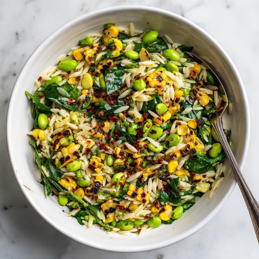 A close-up of a salad served in a white bowl with a speckled brown rim, filled with three layers of fresh ingredients. The bottom layer consists of small, pale yellow orzo pasta mixed with light green edamame beans. The middle layer has bright yellow grilled corn pieces showing slight charring for texture. The top layer is made up of vibrant dark green leafy greens, likely arugula, spread unevenly with some sprinkled herbs and herbs mixed throughout. The bowl is placed on a white marbled surface with a beige cloth partially visible in the background. Photo taken with an iphone --ar 4:5 --v 7